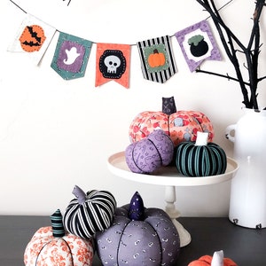 K&ouml;nnte beinhalten: Eine Sammlung handgemachter Halloween-K&uuml;rbisse mit verschiedenen Mustern, darunter Totenk&ouml;pfe, Geister und K&uuml;rbisse. Die K&uuml;rbisse werden auf einem wei&szlig;en Kuchenst&auml;nder und einer grauen Oberfl&auml;che ausgestellt. Ein Halloween-Banner mit verschiedenen Halloween-Symbolen h&auml;ngt &uuml;ber den K&uuml;rbissen.