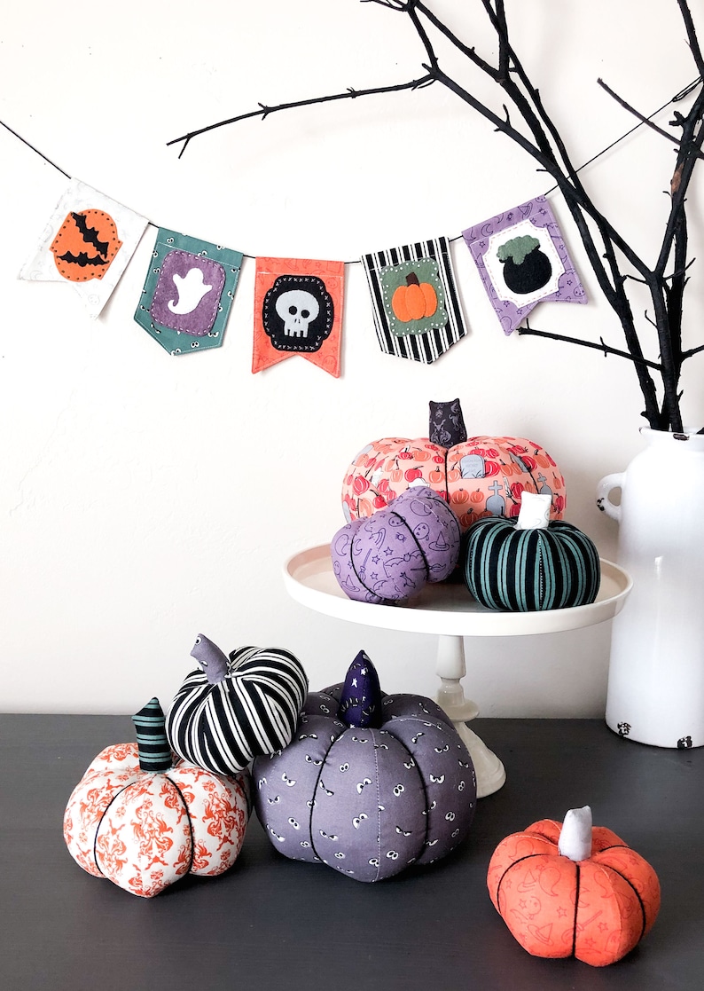 K&ouml;nnte beinhalten: Eine Sammlung handgemachter Halloween-K&uuml;rbisse mit verschiedenen Mustern, darunter Totenk&ouml;pfe, Geister und K&uuml;rbisse. Die K&uuml;rbisse werden auf einem wei&szlig;en Kuchenst&auml;nder und einer grauen Oberfl&auml;che ausgestellt. Ein Halloween-Banner mit verschiedenen Halloween-Symbolen h&auml;ngt &uuml;ber den K&uuml;rbissen.
