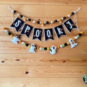 May include: A Halloween-themed felt banner with the word "SPOOKY" spelled out in individual black pennants. The banner is decorated with felt balls in orange, black, white, and green, and includes three white ghost figures. The banner is hanging on a wooden wall.