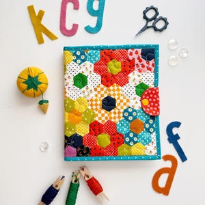 May include: A rectangular fabric cover featuring a colorful patchwork of hexagonal patterns in various prints and colors, secured with a red polka-dot tab. It is surrounded by felt letters, small scissors, buttons, and thread spools.