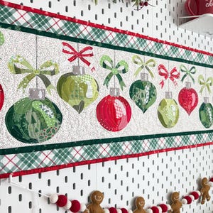 Puede incluir: Un camino de mesa acolchado con un patrón repetitivo de adornos navideños colgantes en tonos rojos y verdes, acentuado con lazos y un borde a cuadros. Debajo, una guirnalda de figuras de pan de jengibre de fieltro y pompones.