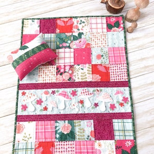 May include: A patchwork quilt with a pink, green and white floral pattern. The quilt is made up of squares of different fabrics, and it has a green border. There are also white felt mushrooms appliqued on the quilt.