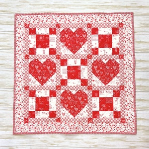 May include: A red and white patchwork quilt with a heart design. The quilt is made of squares with a gingham pattern and a floral print. The quilt is edged with a white and red floral trim.
