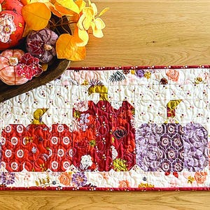 May include: A quilted table runner featuring a pumpkin design in red, orange, and purple. The runner has floral patterns and a red border. A wooden bowl with decorative pumpkins and autumn leaves sits on a wooden surface.