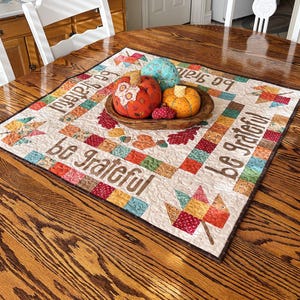 May include: A wooden bowl holds three fabric pumpkins and small red berry decorations, set on a quilted table runner. The runner features a patchwork design with the words "Be Grateful" and leaf motifs. The table is made of wood.