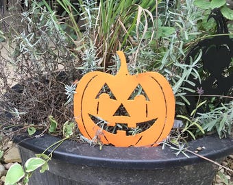 Op de afbeelding: Een platte, oranje metalen pompoenuitsparing met een klassiek jack-o'-lantern-gezicht met driehoekige ogen, een driehoekige neus en een brede, tandige grijns. De pompoen staat rechtop tussen groene planten in een grote, donkergrijze plantenbak.