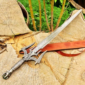 May include: A silver sword with a detailed, intricate design and a leather strap. The sword has a sharp point and a curved handle. The sword is lying on a wooden surface.