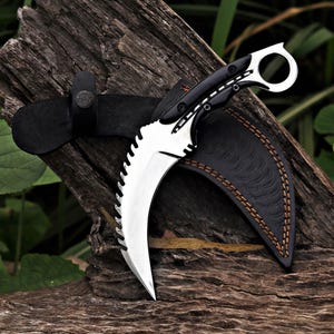 Pode incluir: Uma faca karambit curva com uma lâmina serrilhada e um cabo preto. A faca está colocada numa bainha de couro preta com costuras castanhas. A pega tem um anel para o dedo. A faca é exibida numa peça de madeira envelhecida.