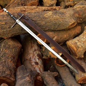 May include: A long, silver sword with a black handle and crossguard, lying on a pile of wooden logs. The sword has a brown leather sheath. The blade is sharp and the handle is detailed.