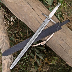 May include: A long, silver and white sword with a patterned blade, resting on a wooden surface. The sword has a white handle and a black leather sheath with a tan strap. The blade has a detailed pattern.