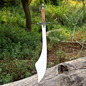 May include: A large, curved sword with a polished steel blade and a wooden handle. The hilt features a decorative cross-shaped guard. The sword is displayed outdoors, resting on a weathered log, with a backdrop of green foliage.