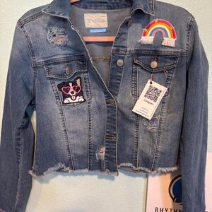May include: A blue denim jacket with distressed details and patches. The patches include a rainbow, a dog wearing heart-shaped sunglasses, and a tag with a QR code. The jacket has a frayed hem.