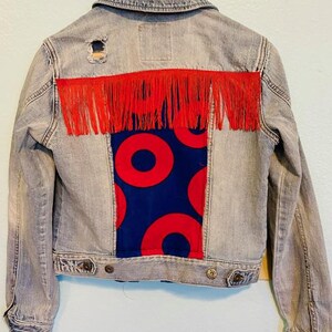 May include: A light gray denim jacket with red fringe and a blue and red fabric patch on the back. The patch features a repeating pattern of red circles on a blue background.