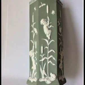 May include: A sage green, octagonal vase with white bas-relief figures and botanical elements. The vase features cherubs, birds, and cattails. The base and rim have decorative detailing. The overall design is classical and elegant.