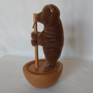 May include: A brown teddy bear figurine holding onto a wooden pole, standing on a wooden bowl-shaped base. The bear has a textured, plush appearance. The base and pole are made of light-colored wood.