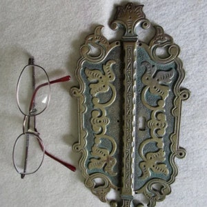 May include: An antique brass door handle with an ornate design, featuring floral and scrollwork details. The handle has a keyhole and is set against a light background. A pair of eyeglasses with red frames rests nearby.