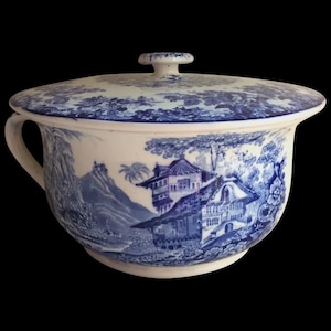 May include: A white and blue ceramic chamber pot with a lid. The pot features a blue and white landscape scene with buildings, trees, and mountains. It has a handle on the side and a small knob on the lid.