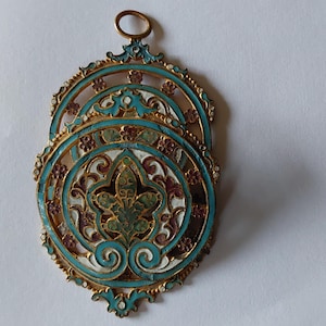 May include: A decorative metal pendant featuring an intricate openwork design with turquoise, white, and dark red enamel accents. The piece has a central floral motif, a circular frame with small flower details, and a small loop at the top for hanging.