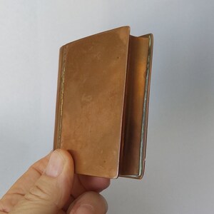 May include: A small, copper-coloured metal book with a hinged spine. The book is open, revealing the inside pages. The edges of the book are slightly rounded. The book is held in a hand against a white background.