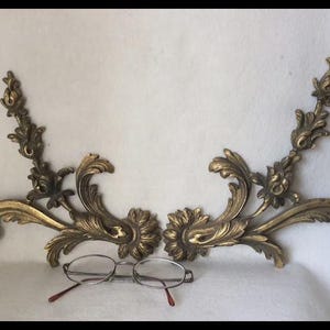 May include: A pair of symmetrical gold-toned metal wall appliques featuring intricate acanthus leaf and floral scrollwork designs. The decorative pieces are positioned above a pair of wire-rimmed eyeglasses with thin metal frames and dark temple tips.