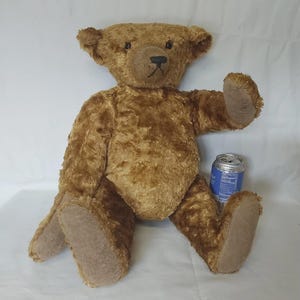 May include: A golden-brown plush teddy bear with textured fur, dark eyes, and a stitched nose. The bear has light tan felt-like fabric on its paw pads and ears. A standard 12-ounce aluminum soda can is positioned next to the bear for scale.