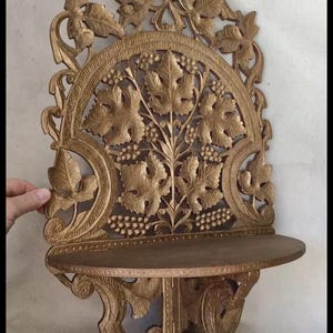 May include: Ornate, golden-colored wooden wall shelf with a carved grape vine design. The shelf has a semi-circular top with a small, rounded shelf. The carved details include grape leaves and bunches of grapes.