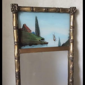 May include: A decorative mirror with a gold-colored bamboo-style frame. The top section features a painted landscape with a house, trees, and a sailboat on a blue background. The lower section is a reflective mirror surface.