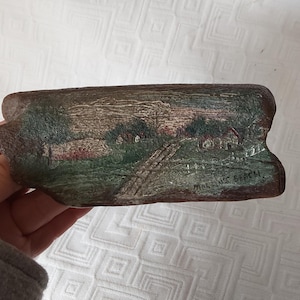 May include: A rustic, rectangular, hand-painted stone featuring a landscape scene with a house, trees, and a path. The artwork is in muted tones of green, brown, and red. The artist's signature, "MARTINE BEACH", is visible.