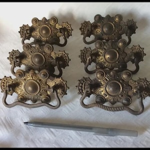 May include: Six antique brass drawer pulls with ornate floral and circular designs. Each pull has a central circular element and a curved handle. The pulls are arranged on a white surface, with a pen in the foreground.