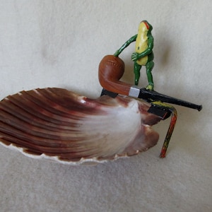 May include: A decorative seashell ashtray with a brown and white interior. A small, green frog figurine stands on a pipe, with a black and red metal stand. The pipe is brown with a silver band.