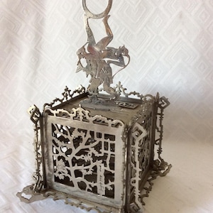 May include: An antique silver-toned metal box with intricate cut-out designs. The box features a decorative figure on top, holding a ring. The sides of the box have a tree-like pattern. The box is supported by four small feet.