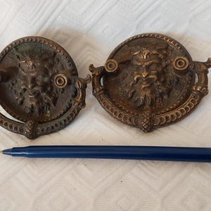 May include: Two circular bronze drawer pulls featuring a relief of a bearded face, each with a swinging drop handle. The pulls are positioned above a dark blue ballpoint pen, which serves as a visual reference for the scale of the hardware.