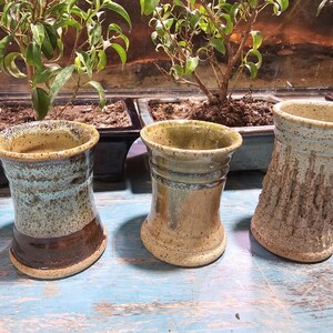 May include: Three handmade ceramic vases in varying earth tones. The vases have a cylindrical shape with textured surfaces. One vase is blue and brown, another is tan, and the third is a light beige with vertical grooves. They are displayed on a blue wooden surface.