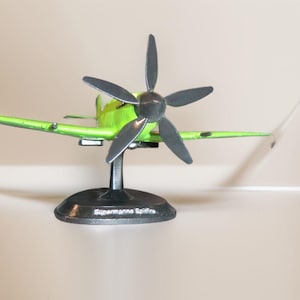 Puede incluir: Un modelo de avión verde y negro en un soporte. El avión tiene una hélice y está etiquetado como "Supermarine Spitfire".