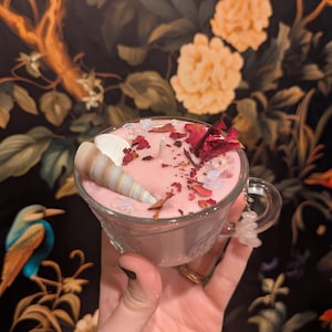 May include: A pink candle in a clear glass teacup, adorned with dried rose petals, small crystals, and a seashell. The candle has a seashell and is surrounded by decorative elements. The teacup is held in a hand.