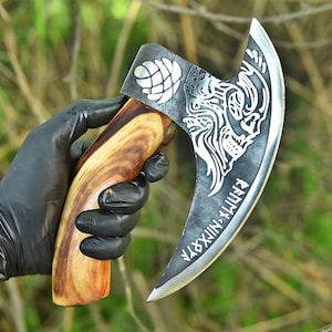 Puede incluir: Un hacha artesanal con una hoja curva y un mango de madera. La hoja presenta un diseño de lobo y runas. El mango es de madera marrón clara y oscura. El hacha está sujeta por una mano enguantada.