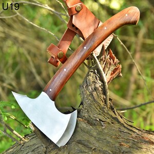 May include: A small, hand-forged axe with a brown wooden handle and a leather sheath. The axe is resting on a tree branch.