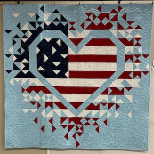 May include: A patriotic quilt featuring a heart-shaped design with the American flag at its center. The quilt showcases red and white stripes, a blue field with white stars, and a light blue background with a geometric pattern. The quilt is a handmade item.
