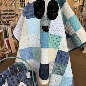 May include: A handmade quilt in shades of blue, white, and green, featuring a patchwork design. The quilt is draped over a display, with large black oval shapes and star details. A basket of fabric bundles and a book are in the foreground.