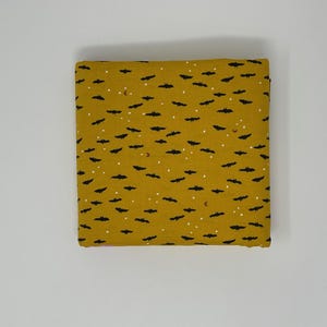 May include: Mustard-colored fabric with a repeating pattern of black bats, white dots, and small crescent moons. The fabric is folded neatly, suggesting it is ready for use in a sewing or crafting project. The design has a Halloween theme.