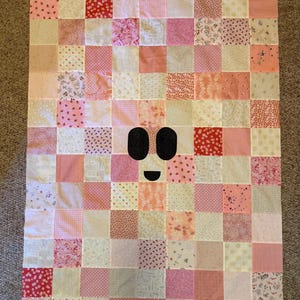 May include: A rectangular patchwork quilt constructed from numerous square fabric patches in various shades of pink, white, and cream. The quilt features a central ghost-like face design composed of two black oval eyes and a small black mouth.
