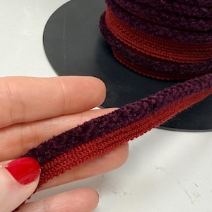 May include: A spool of burgundy and red velvet trim. The trim is a combination of a plush, textured burgundy material and a smooth, red fabric. The trim is rolled up on a black surface, with a length of the trim held in a hand.