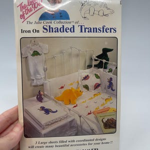 May include: A package of iron-on shaded transfers from The Julie Cook Collection. The package features a bed with dinosaur-themed bedding, a stuffed dinosaur, and the text "Iron On Shaded Transfers".