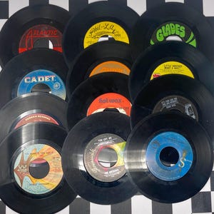 Pode incluir: Uma coleção de discos de vinil vintage de 45 RPM com rótulos coloridos. Os rótulos incluem "Atlantic", "Cadet", "Phil-La of Soul", "Glades" e "Hot Wax". Os discos estão dispostos numa superfície quadriculada a preto e branco.