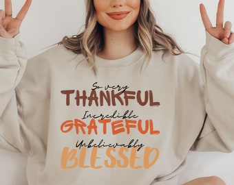 May include: A beige sweatshirt with the text "So very Thankful Incredible Grateful Unbelievably Blessed" in brown, orange, and yellow lettering.