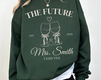 May include: Forest green sweatshirt with the text "THE FUTURE" and "Mrs. Smith I SAID YES!". The design includes two wine glasses, a heart, and a ring. The year "2026" is also printed on the sweatshirt.