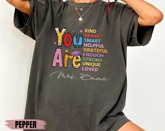 May include: A dark gray t-shirt with the words "You Are" in colorful letters. The words "Kind, Brave, Smart, Helpful, Grateful, Enough, Strong, Unique, Loved" are stacked vertically in rainbow colors. The text "Mrs. Emma" is written in white below the words.