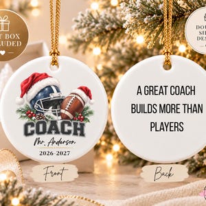 May include: A round white ceramic ornament with a gold hanging cord. The front features a football helmet and football wearing Santa hats, the text COACH, Mr. Anderson, and 2026-2027. The back displays the text A GREAT COACH BUILDS MORE THAN PLAYERS.