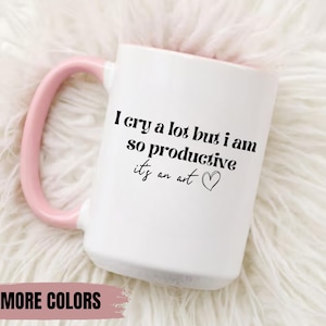 May include: White ceramic mug with a pink handle. The mug has black text that reads "I cry a lot but I am so productive it's an art" with a small heart symbol.