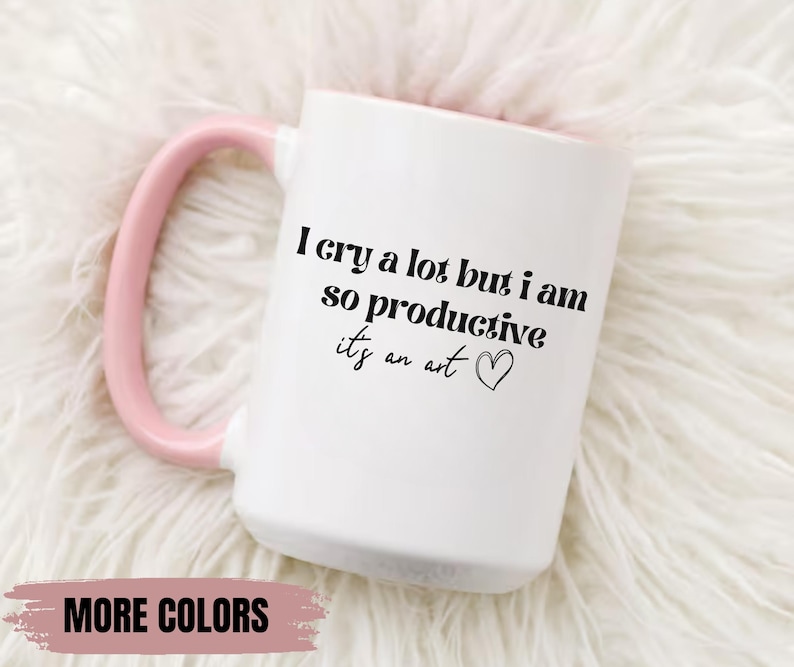 May include: White ceramic mug with a pink handle. The mug has black text that reads "I cry a lot but I am so productive it's an art" with a small heart symbol.